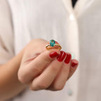 Hand wearing a gold ring with a paraiba gemstone against a neutral background