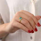 Hand wearing a gold ring with a green gemstone against a neutral background