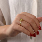 Hand wearing a gold ring with a heart-shaped gemstone against a neutral background