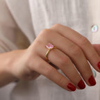 Hand wearing a gold ring with a pink gemstone against a neutral background