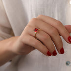 Hand wearing a gold ring with a red gemstone on a neutral background