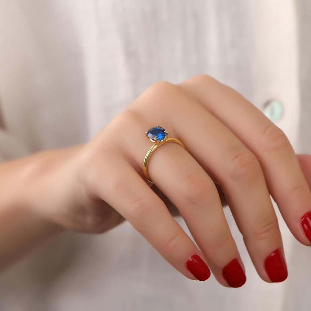 Hand wearing a gold ring with a blue gemstone on a plain background