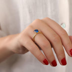Hand wearing a gold ring with a blue gemstone on a plain background
