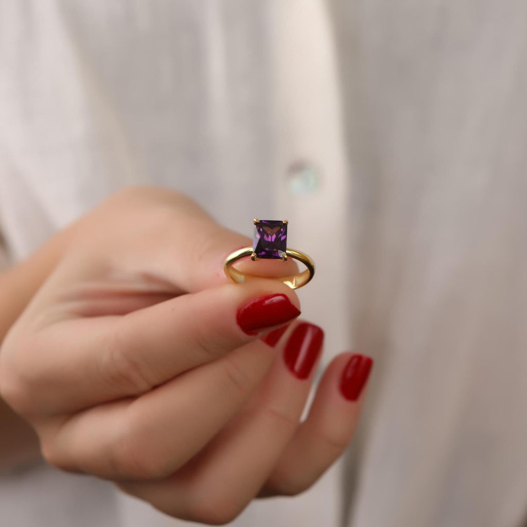 Gold ring with a purple gemstone held by a hand with red nail polish against a neutral background