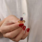 Gold ring with a purple gemstone held by a hand with red nail polish against a neutral background