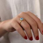 Hand wearing a solitaire ring with a blue gemstone against a neutral background