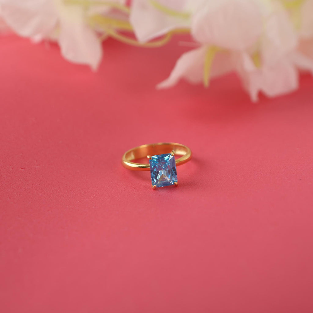 Gold ring with a radiant cut blue gemstone on a pink background with flowers