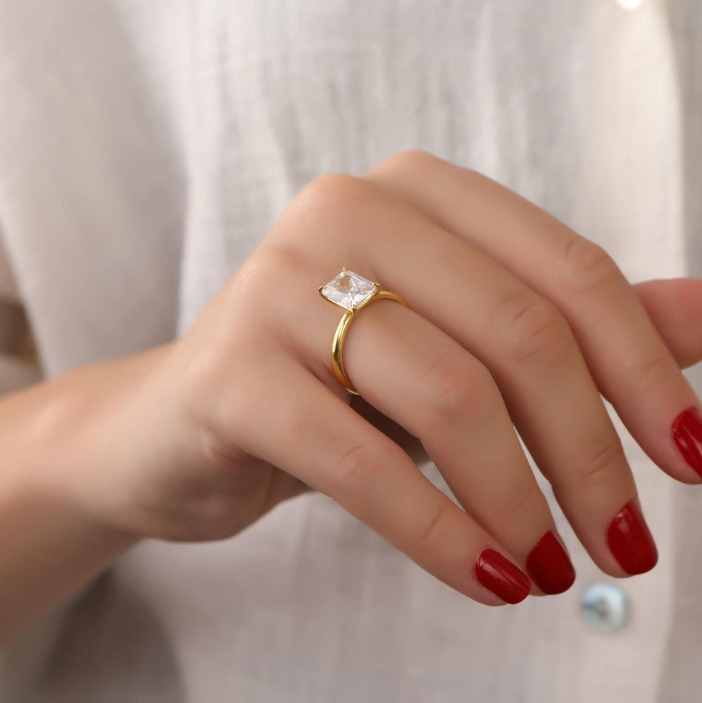 Hand wearing a gold ring with a clear gemstone against a neutral background