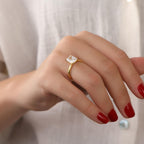 Hand wearing a gold ring with a clear gemstone against a neutral background