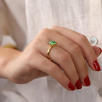 Hand wearing a gold ring with a green emerald gemstone against a neutral background