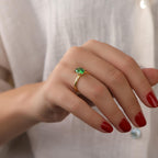 Hand wearing a gold ring with a green gemstone against a neutral background