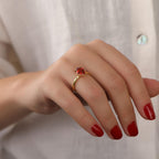 Hand wearing a gold ring with a red gemstone against a neutral background