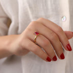 Hand wearing a gold ring with a red gemstone against a neutral background