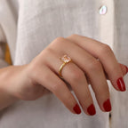 Hand wearing a gold ring with a pink gemstone against a neutral background