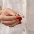 Hand with red nail polish holding a gold ring with a morganite gemstone against a blurred background