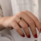 Hand wearing a gold ring with a black gemstone against a neutral background