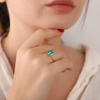 Close-up of a hand wearing a gold ring with a green gemstone paraiba, touching the chin.