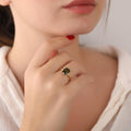 Close-up of a hand wearing a gold ring with a green gemstone, touching the chin.