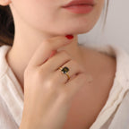 Close-up of a hand wearing a gold ring with a green gemstone, touching the chin.