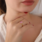 Close-up of a hand wearing a gold ring with a pink gemstone, blurred background