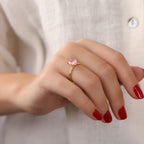 Hand wearing a gold ring with a pink gemstone against a neutral background