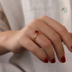 Hand wearing a gold ring with a red gemstone against a neutral background