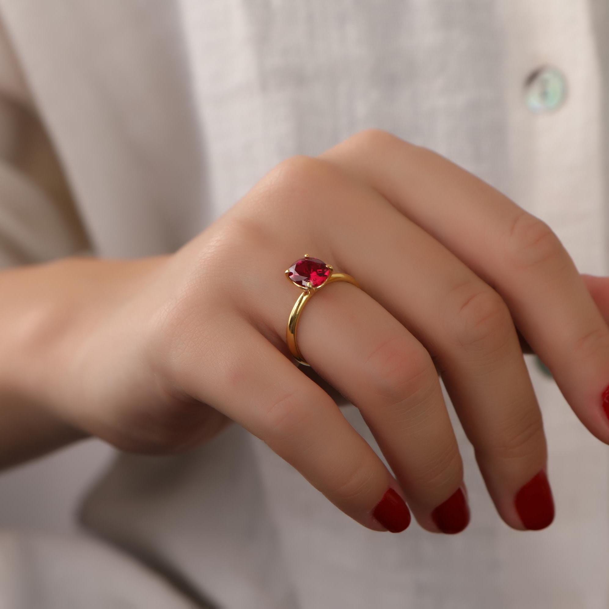 Hand wearing a gold ring with a red gemstone against a neutral background