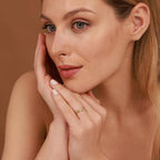 Woman with a soft smile, gently touching her face with a twist ring against a warm brown background