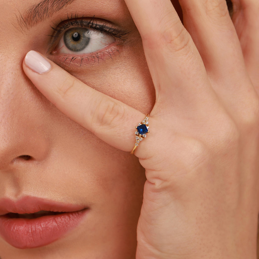 Close-up of a woman wearing a gold ring with a blue gemstone, holding her hand to her face.