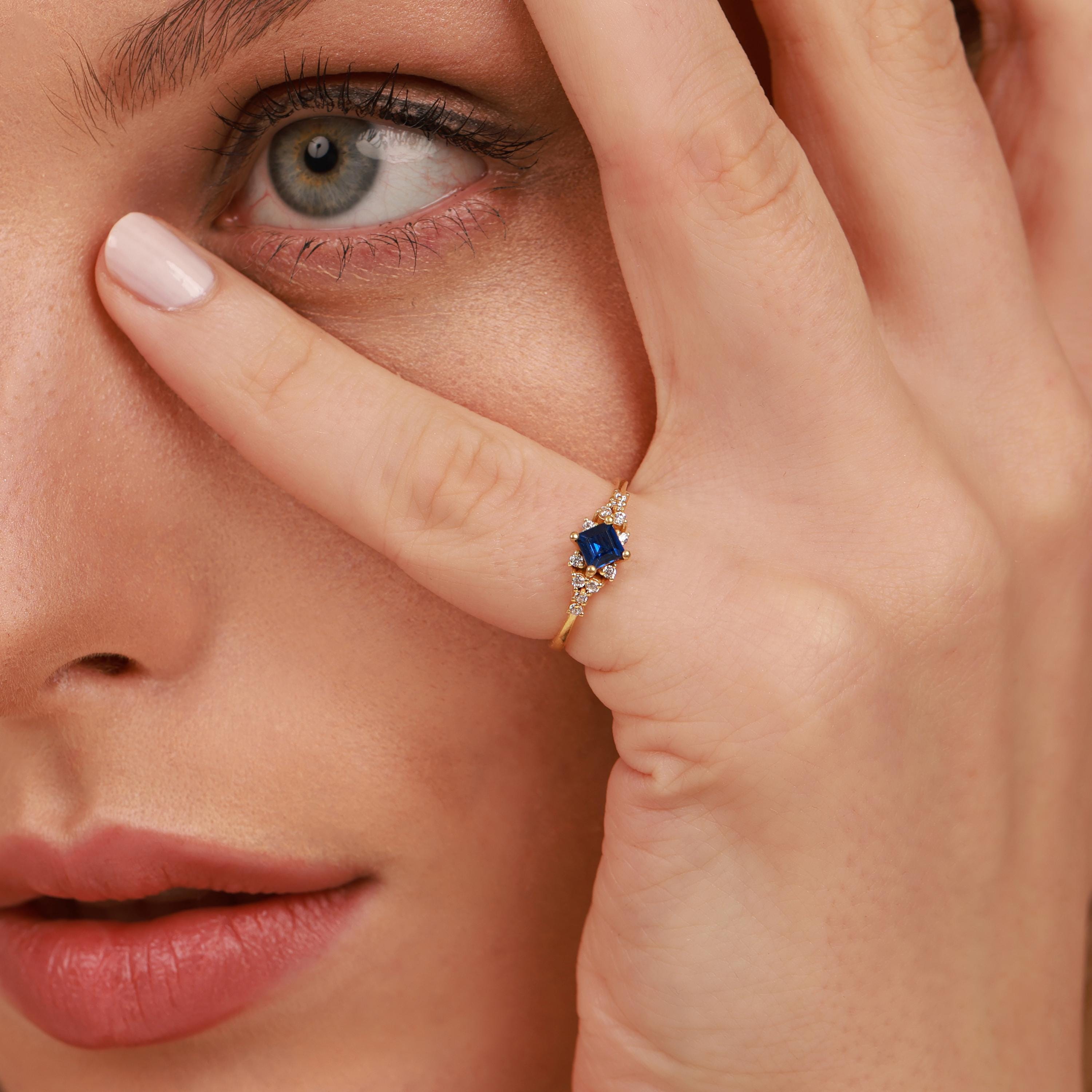 Close-up of a woman wearing a gold ring with a blue gemstone, holding her hand to her face.