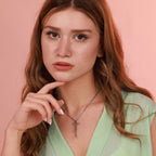 May include: A silver-toned crucifix pendant necklace. The detailed pendant depicts a figure on a cross. The necklace is worn against a light green top and a pink background. The chain is a delicate, linked design.