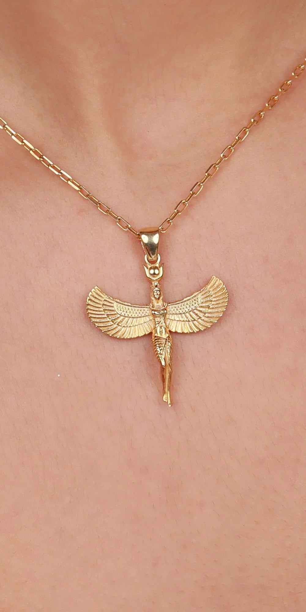 A gold-toned necklace with a winged pendant featuring a small figure in the center, shown worn on a person's neck against a light skin tone background.