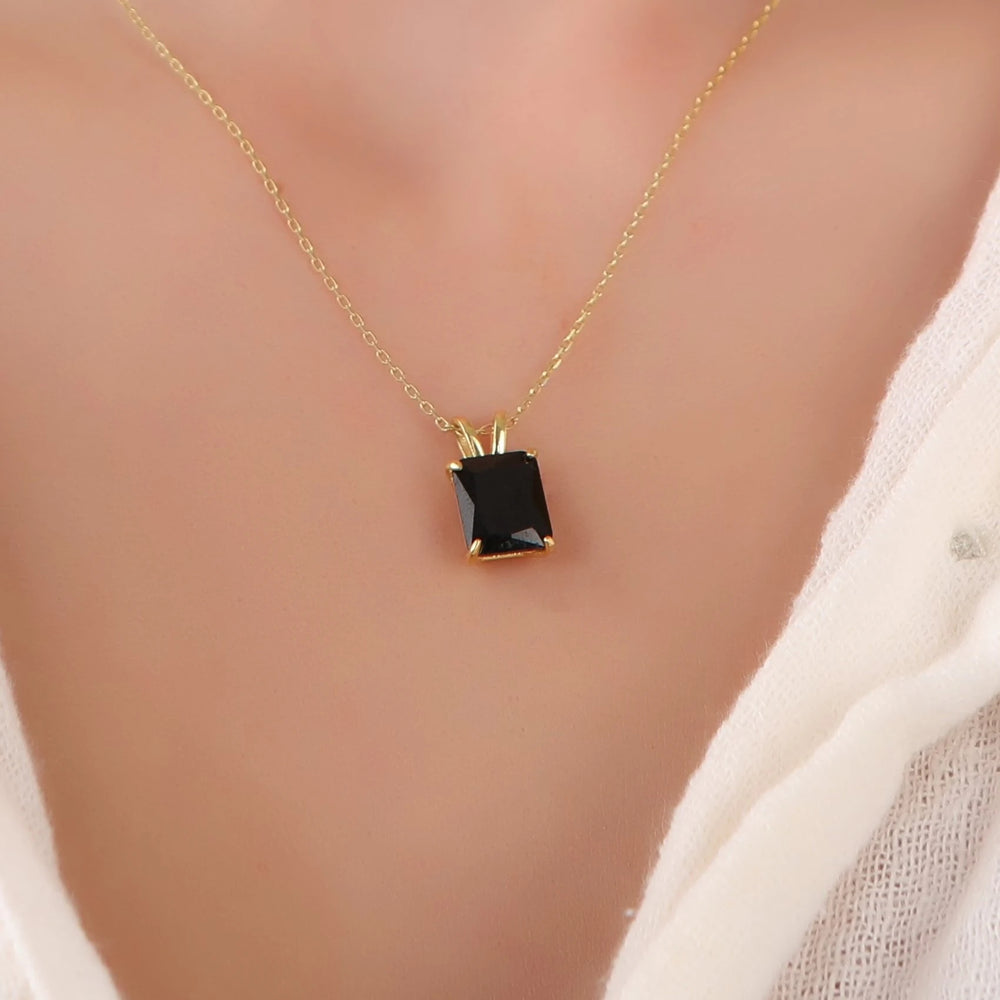 A close-up of a person's chest wearing a delicate gold chain necklace with a rectangular black gemstone pendant in a gold setting, paired with a white knit top.