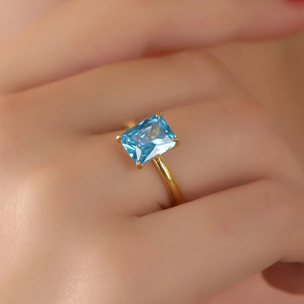 A gold ring with a rectangular light blue gemstone worn on a person's finger, shown in a close-up shot.