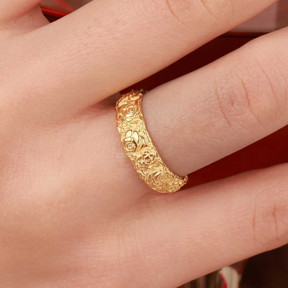 A close-up of a hand wearing a textured yellow gold ring with an intricate floral or rose pattern, set against a soft red background.