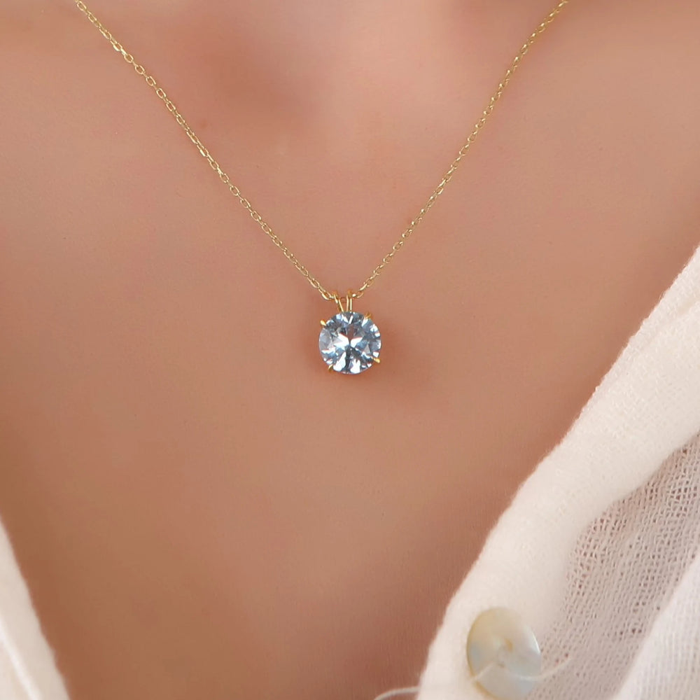 A delicate gold chain necklace with a single round clear gemstone pendant, worn on a model's neck over a white knit top.