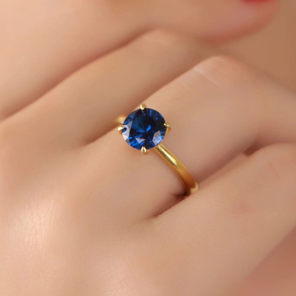 A gold ring with a round-cut deep blue gemstone, shown worn on a person's finger.