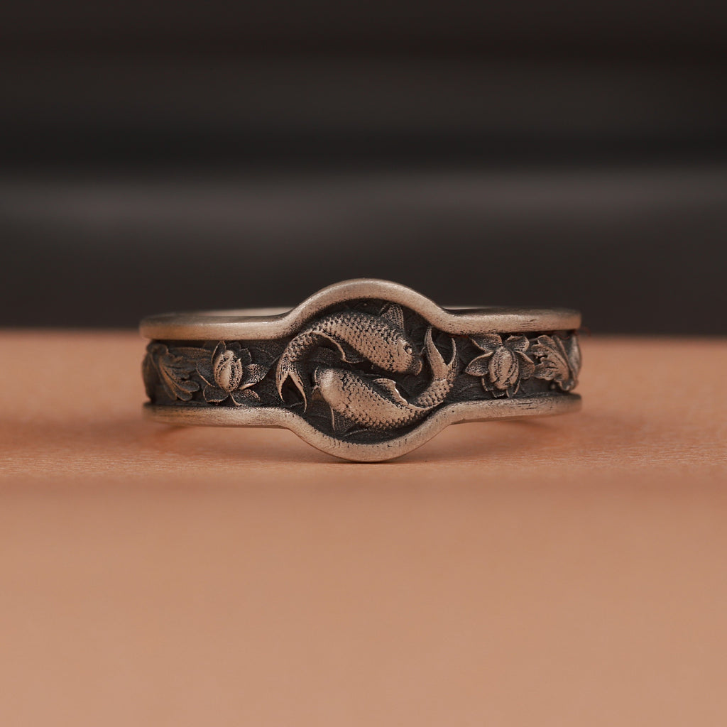 Silver ring with fish and lotus designs on a brown background