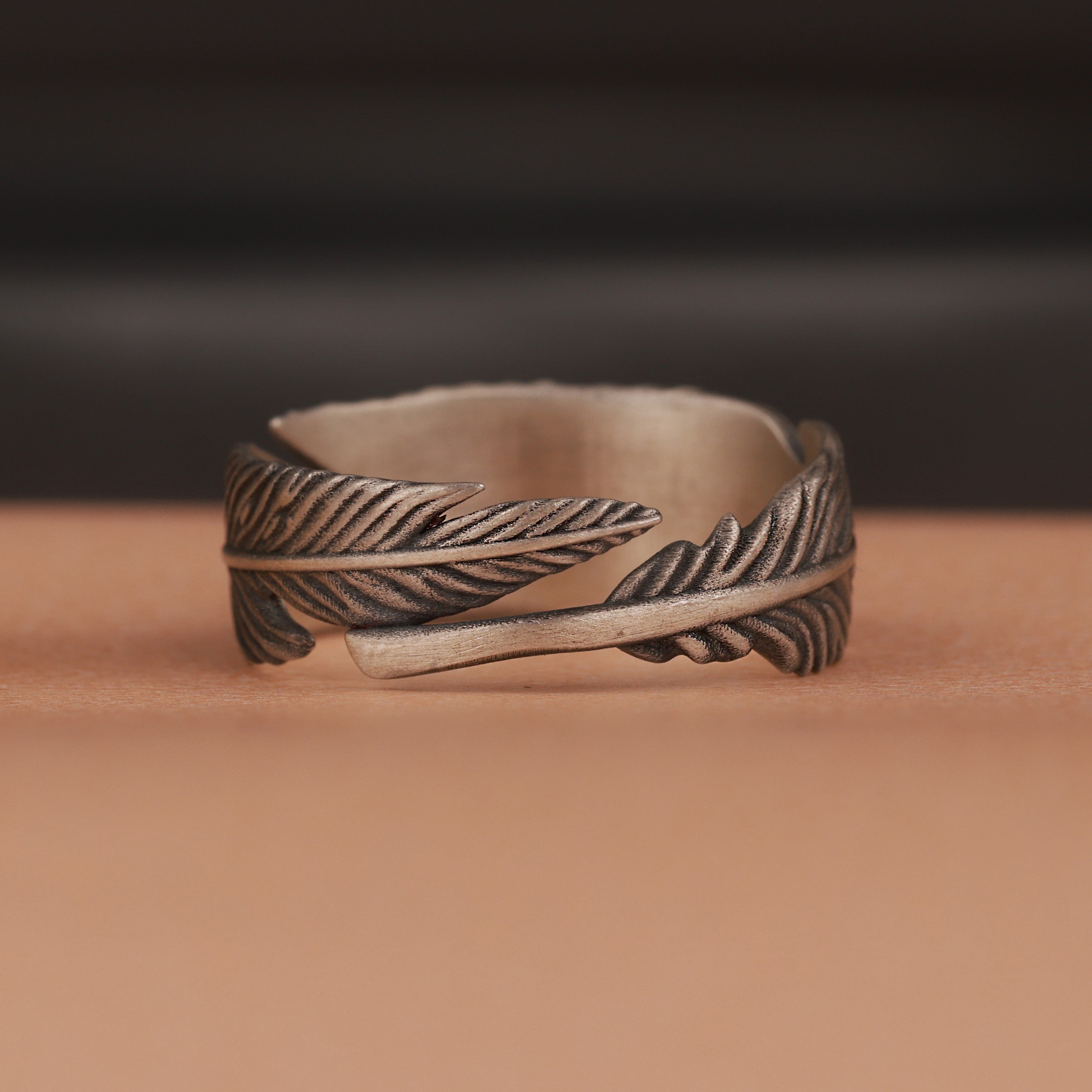 Oxidized silver feather-shaped ring on a brown surface with a blurred dark background